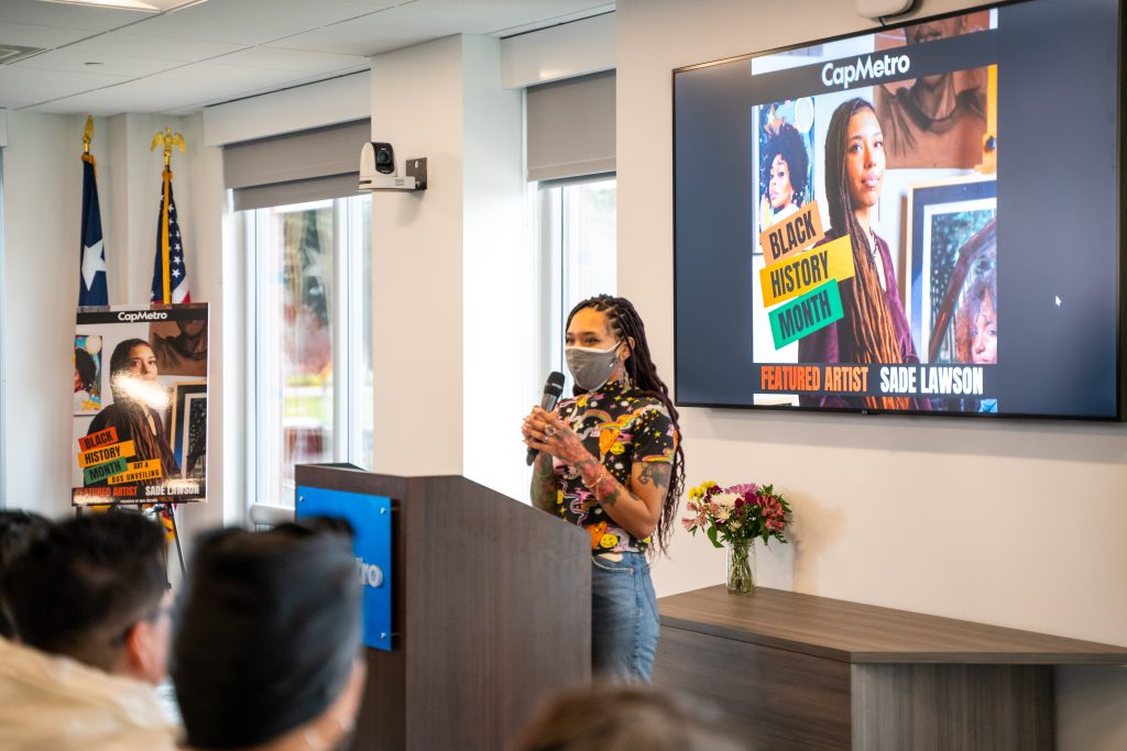 Photo of artist Sadé Lawson holding a microphone and speaking in front of a podium inside.